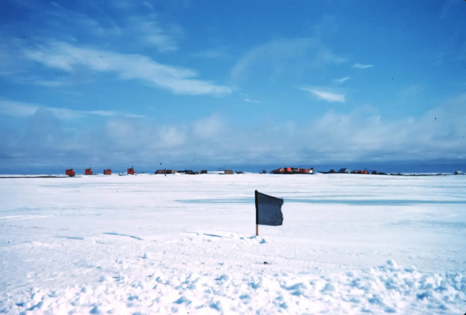 A flag blown straight out on its pole above a snow-covered airstrip at Oliktok Point, Alaska, under a blue sky.